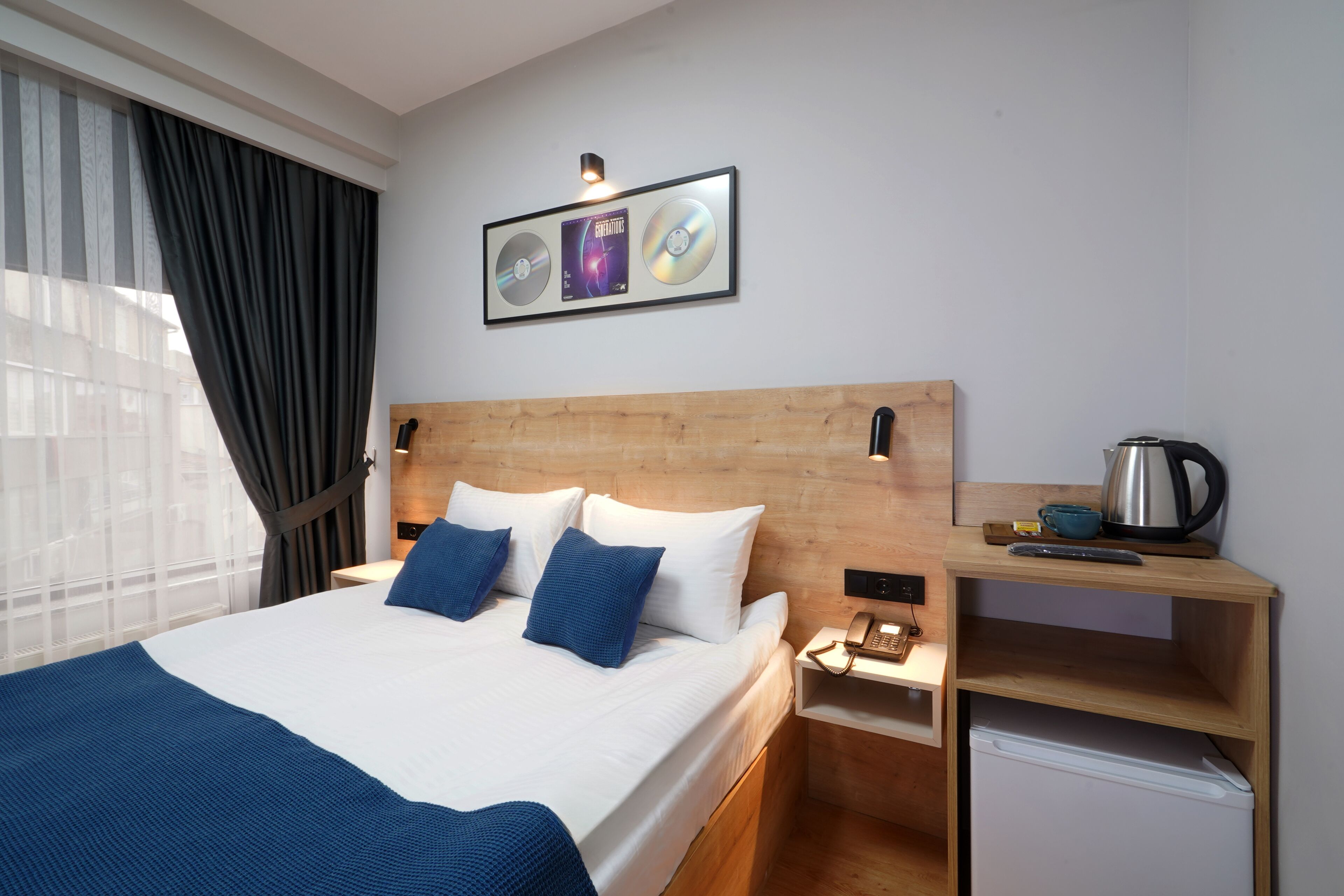 A hotel room with a wooden headboard, a bed with white and blue bedding, a small desk with a kettle, and a framed picture of CDs on the wall.