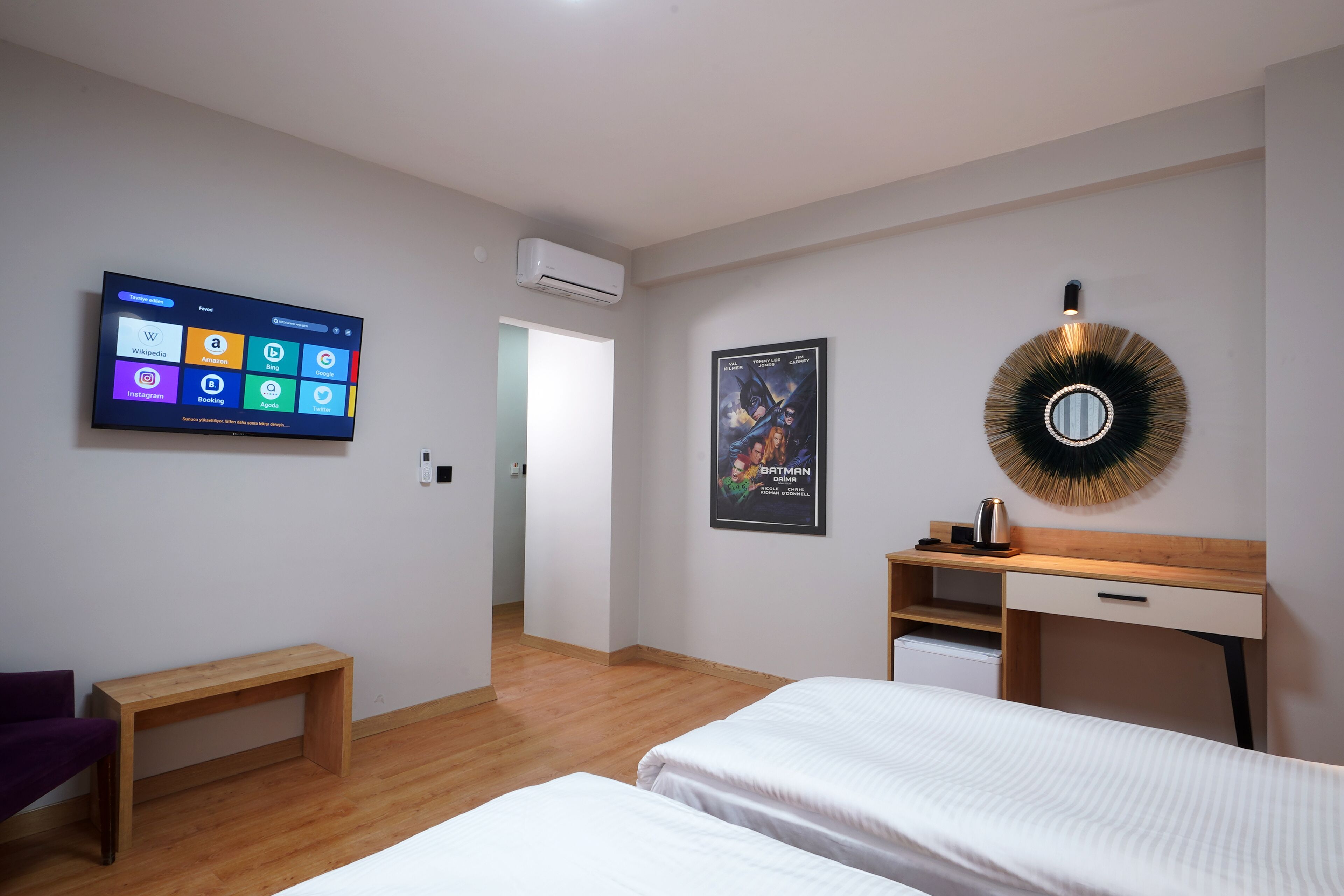 Una habitación de hotel con una televisión de pantalla plana mostrando la interfaz de una aplicación de redes sociales, un cartel de Batman, un espejo redondo, un escritorio de madera y dos camas.