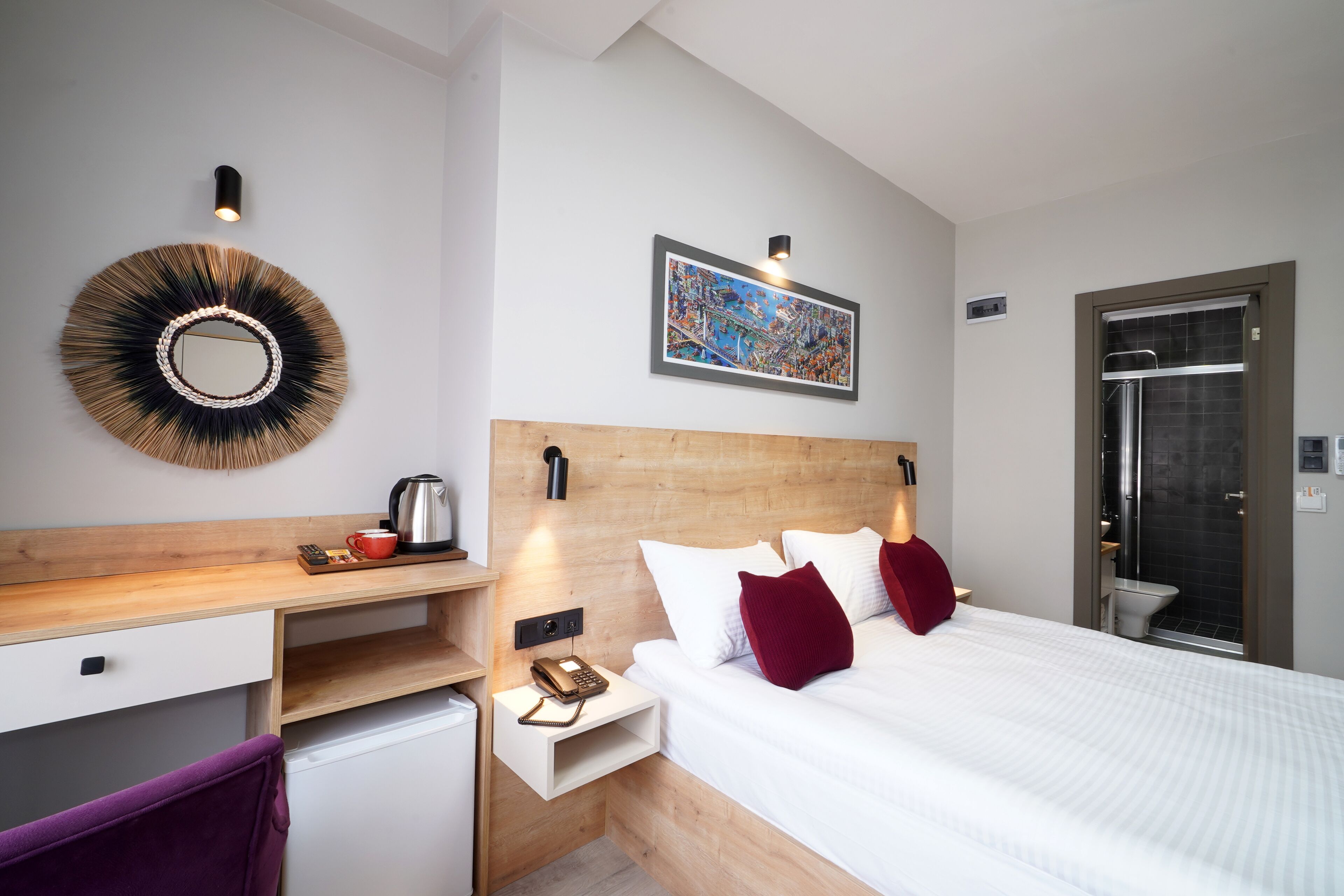 Una camera d'albergo moderna con una testiera in legno, un letto con biancheria da letto bianca e cuscini rossi, una piccola scrivania con una macchina per il caffè, uno specchio rotondo e un quadro con una vista sulla città.