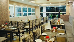 A restaurant with a long dining table, multiple chairs, and a sofa. There are large windows and a ceiling fan. -  The Century Stays (Jaipur)