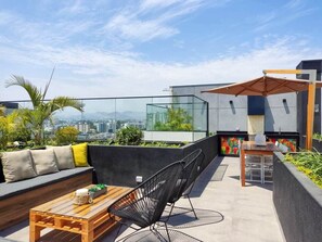 Terraço no telhado com guarda-corpo de vidro, mesa de madeira, sofá com almofadas, uma cadeira e uma pequena mesa com uma planta.