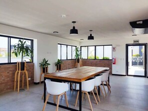 A spacious room with a large wooden table and white chairs, large windows, and potted plants.