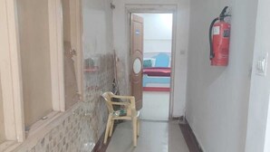 A hallway with a tiled floor, a fire extinguisher mounted on the wall, and a door leading to a room with a bed. - Hotel Abhishek Residency By The Arrow (New Delhi)