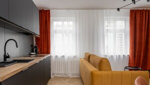 Et moderne køkken med træbordplade, indbygget komfur og vask. I opholdsstuen er der en sofa og en lænestol, samt et vindue med gardiner.