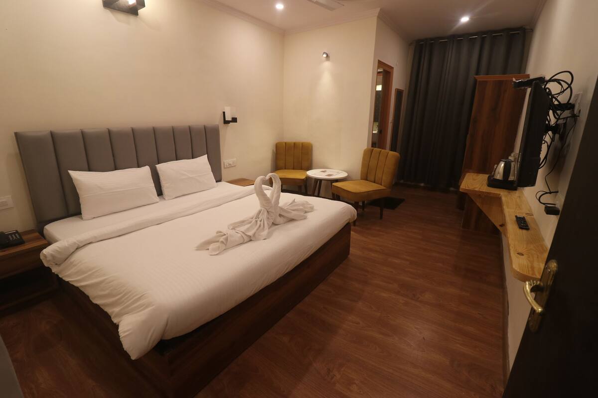 A hotel room with a large bed, a small table, two chairs, a desk with a kettle, and a TV.