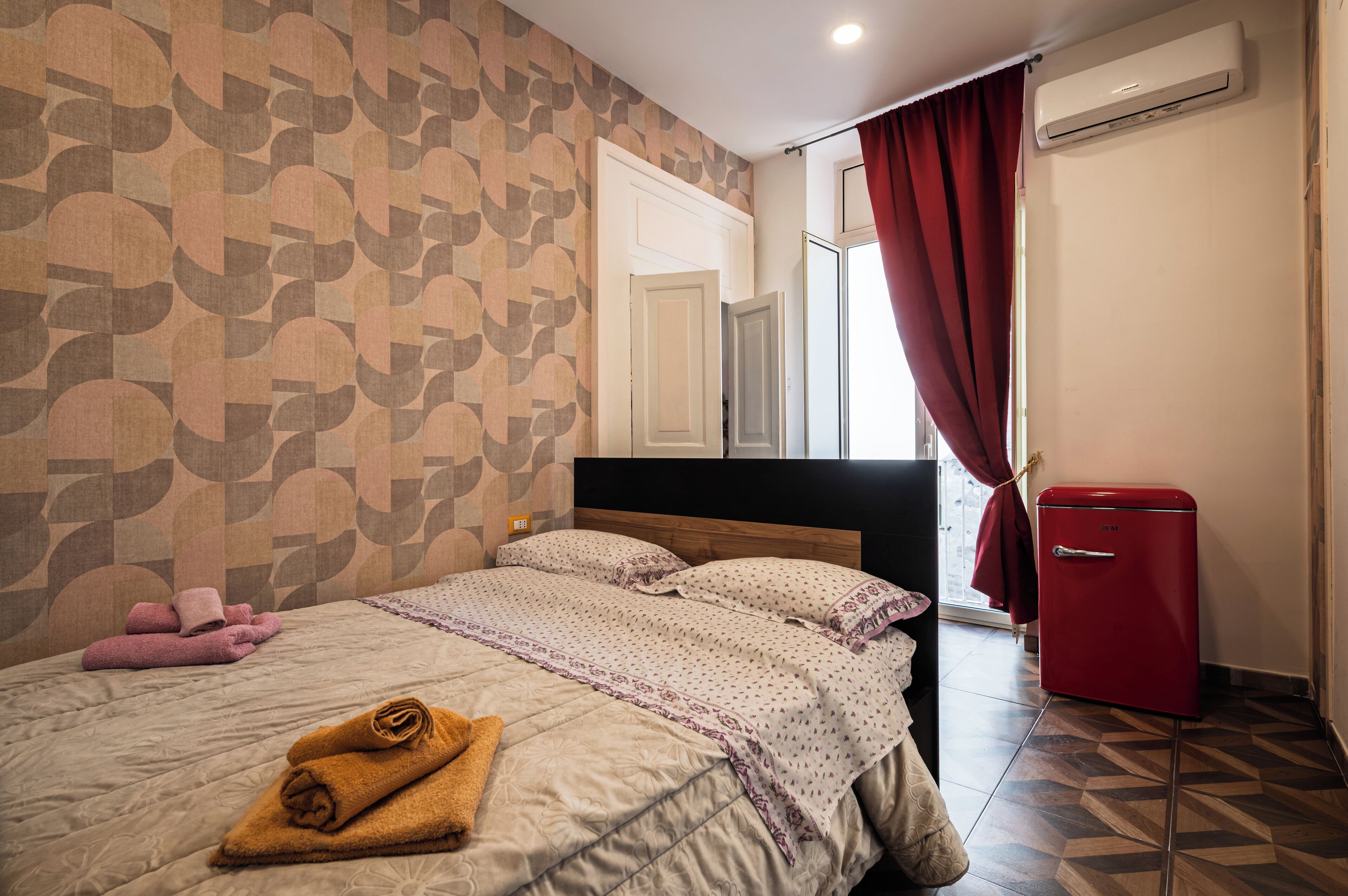 A neatly made bed with rolled towels, a red refrigerator, and a patterned wall.