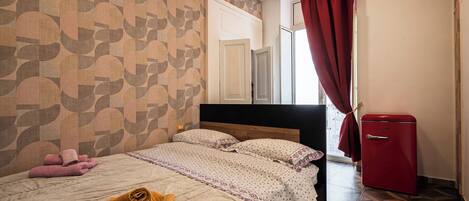 A neatly made bed with rolled towels, a red refrigerator, and a patterned wall.