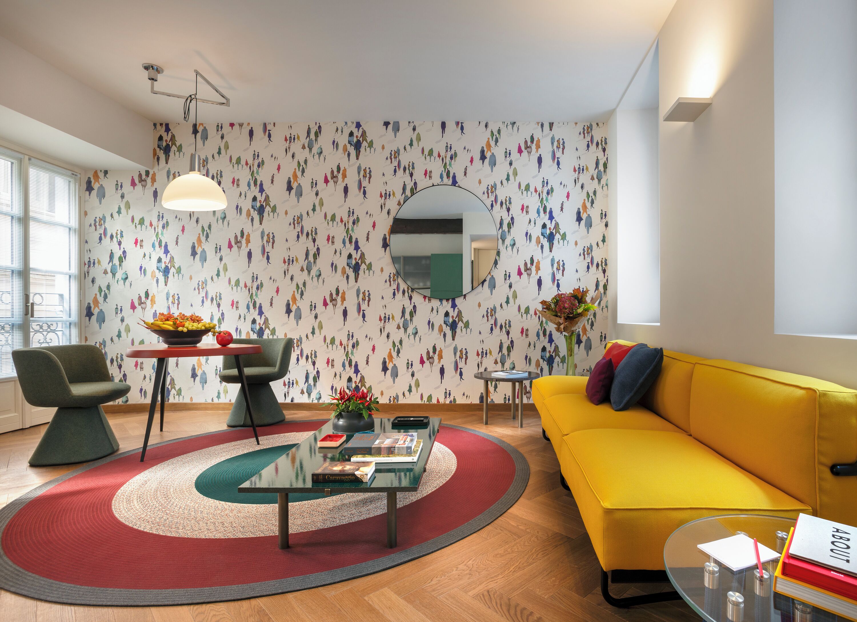 A modern living room with a yellow sofa, a round glass coffee table, a green armchair, a red and green rug, and a wall with a colorful abstract pattern.