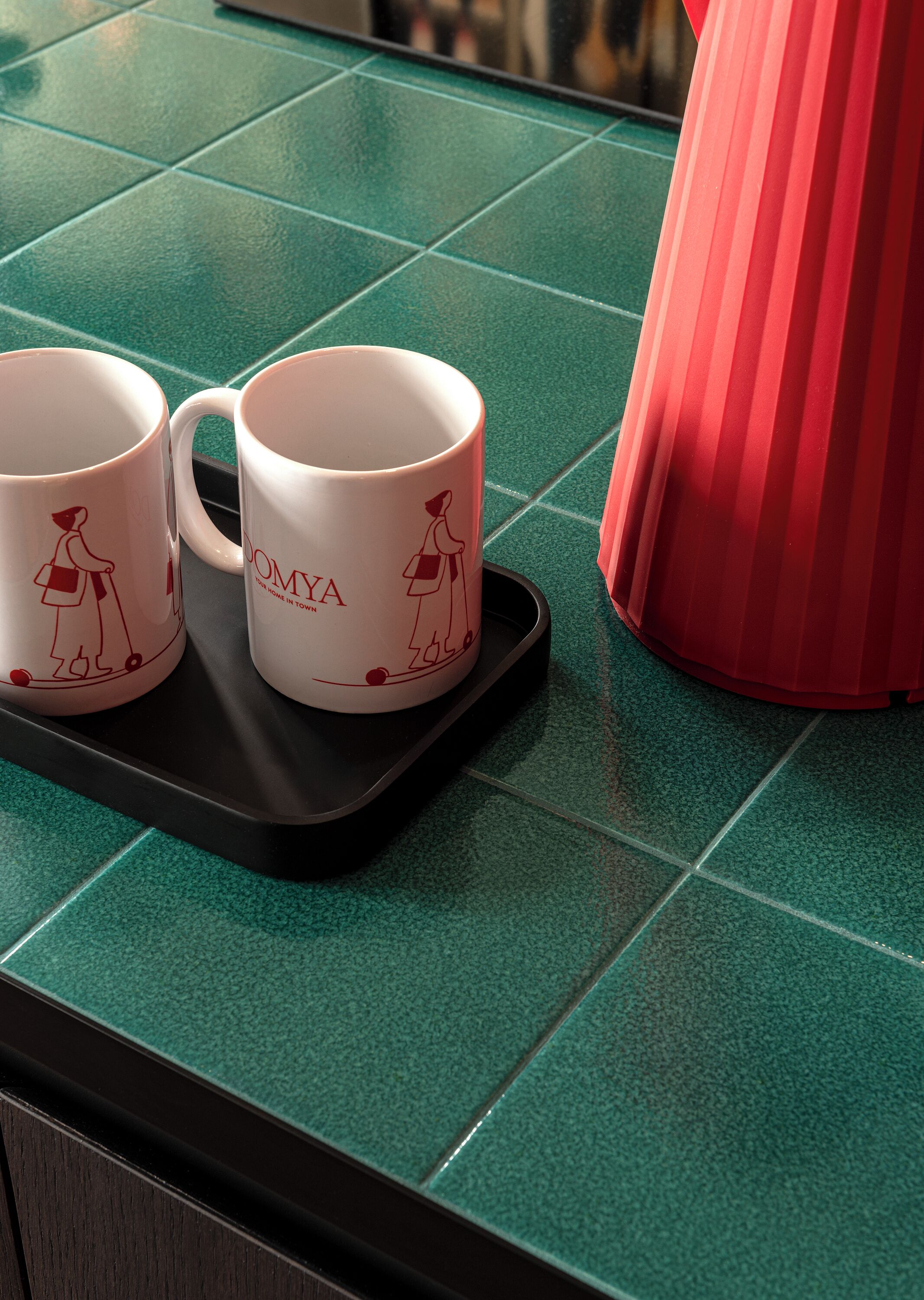 Two white mugs with red designs on a black tray on a green tiled countertop.