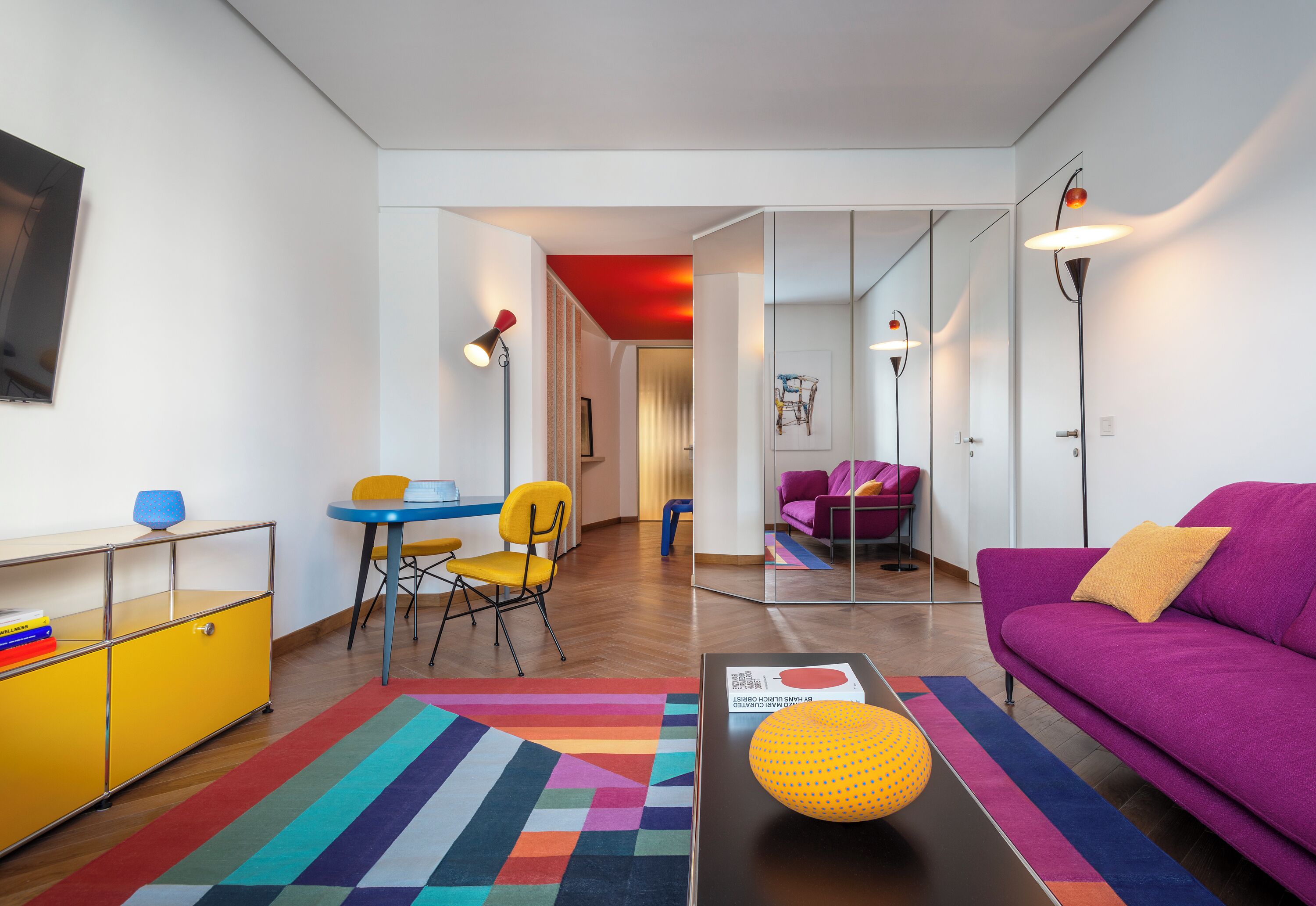 A modern living room with a purple sofa, a yellow chair, a small table with a lamp, and a colorful rug.