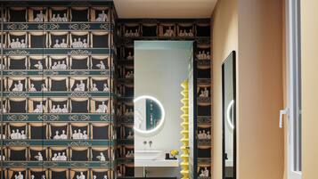 Une salle de bain moderne avec un grand miroir, un lavabo et des toilettes. On y trouve également un ballon d’exercice noir et une valise posés sur un banc.