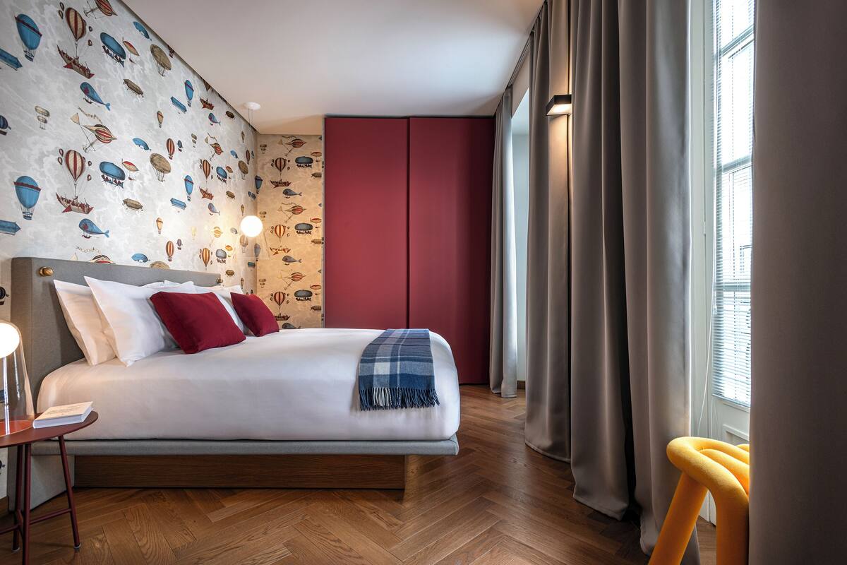 Une chambre moderne avec un papier peint à motifs, une armoire rouge, un lit avec une literie blanche et des oreillers rouges, un plaid bleu et un plancher en bois.