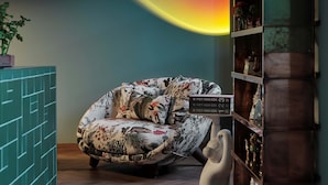 A room with a colorful light installation on the wall, a patterned sofa, a white chair, and a wooden bookshelf with decorative items. - DOMYA - Manzoni 25 (Milan)