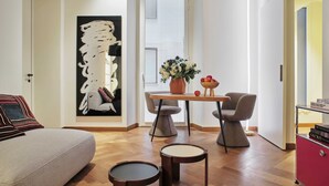 A modern living room with a sofa, a coffee table, and two chairs. There is a large abstract painting on the wall, a potted plant, and a small table with fruits and books. - DOMYA - Manzoni 25 (Milan)
