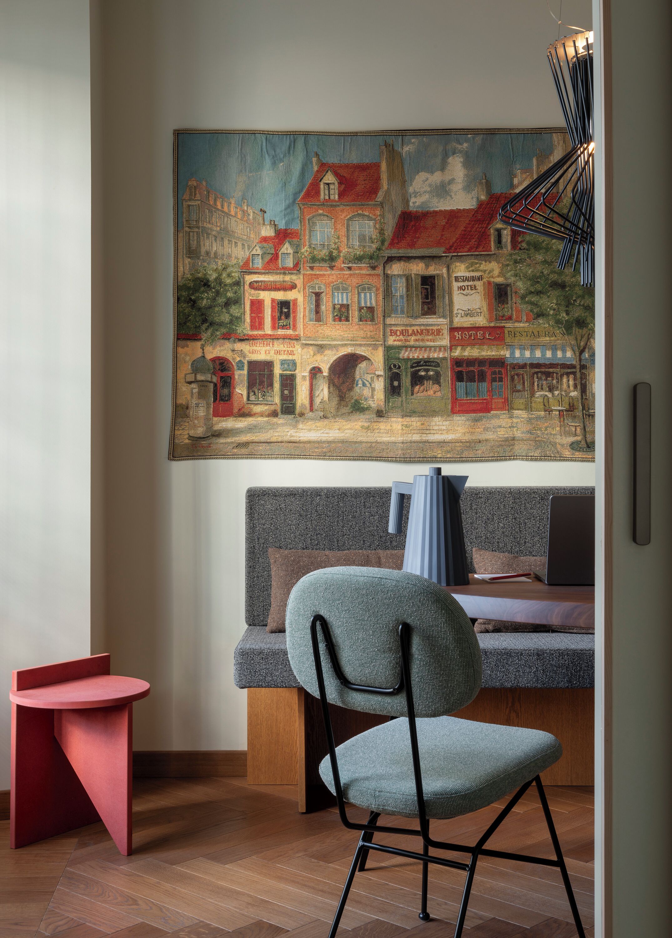A cozy room with a large painting of a European street scene, a modern sofa, a small red table, and a chair with a blue cushion.