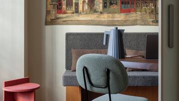 A cozy room with a large painting of a European street scene, a modern sofa, a small red table, and a chair with a blue cushion.