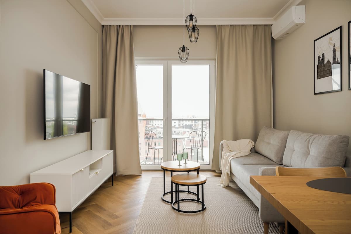 A modern living room with a flat-screen TV, a sofa, a wooden table, and a balcony with a view of buildings.