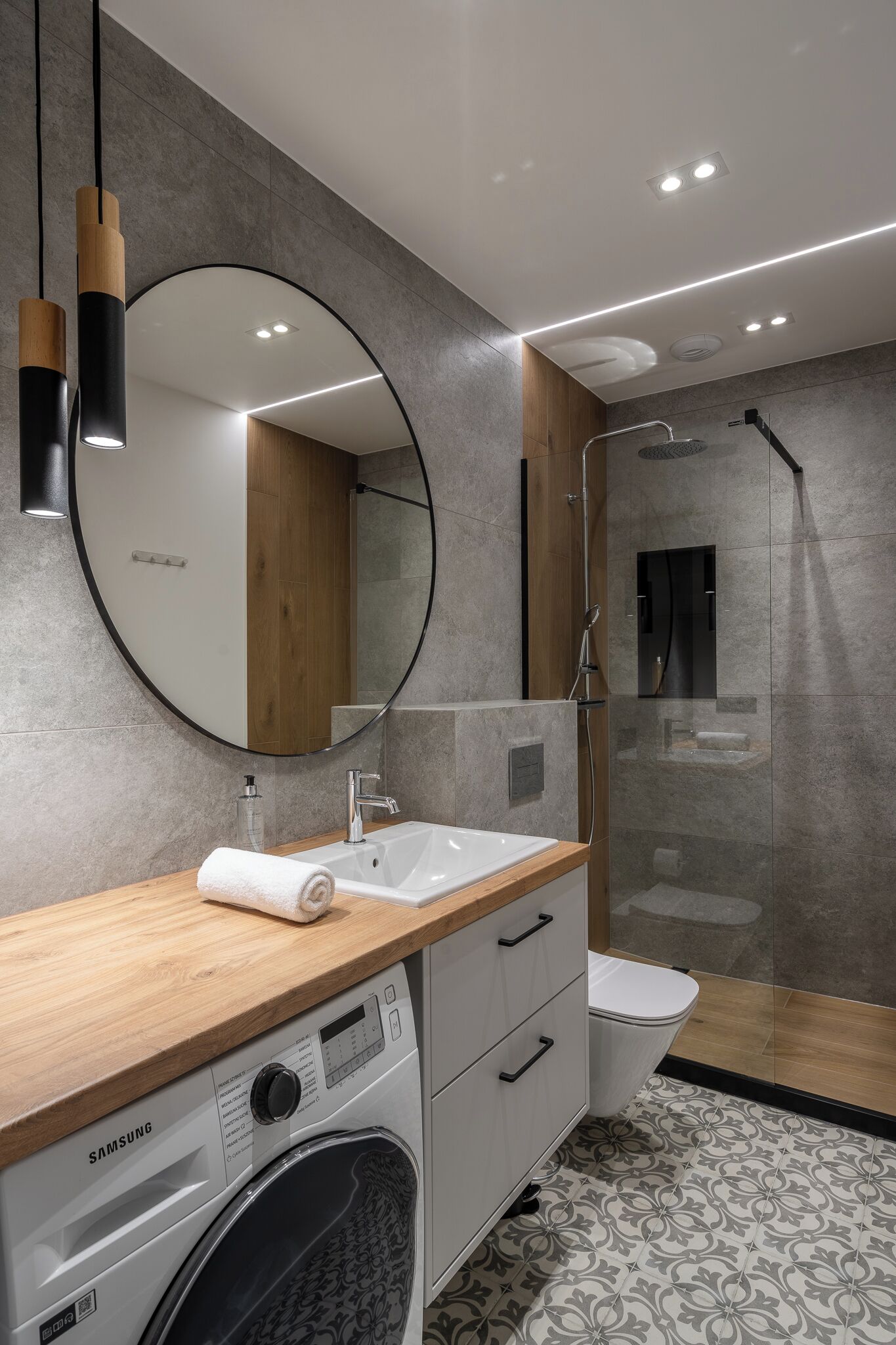 A modern bathroom with a wooden vanity, a round mirror, a built-in washing machine, a toilet, and a glass-enclosed shower area.