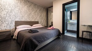 Camera da letto moderna con un letto grande, pavimento in legno scuro, carta da parati decorata e bagno visibile attraverso una porta aperta.