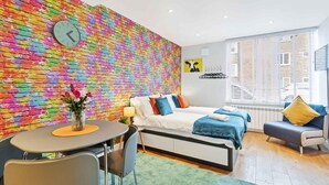A modern living room with a colorful brick wall, a sofa set, a dining table with chairs, and a large window with blinds. - Rainbow Studio - Spacious City Centre - Close to sea - Sleeps 1 to 3 Guests (Brighton)