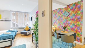 A hotel room with a bed, a seating area with a table and chairs, a colorful brick wall, and a window with blinds. - Rainbow Studio - Spacious City Centre - Close to sea - Sleeps 1 to 3 Guests (Brighton)