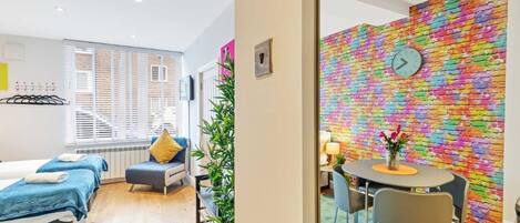 Une chambre d’hôtel avec un lit, un coin salon comprenant une table et des chaises, un mur en briques aux couleurs vives et une fenêtre avec des stores.