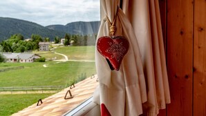 A window with curtains, a heart-shaped decoration, and a view of a green landscape with houses and mountains. - RESIDENCE STELLA (Folgaria)