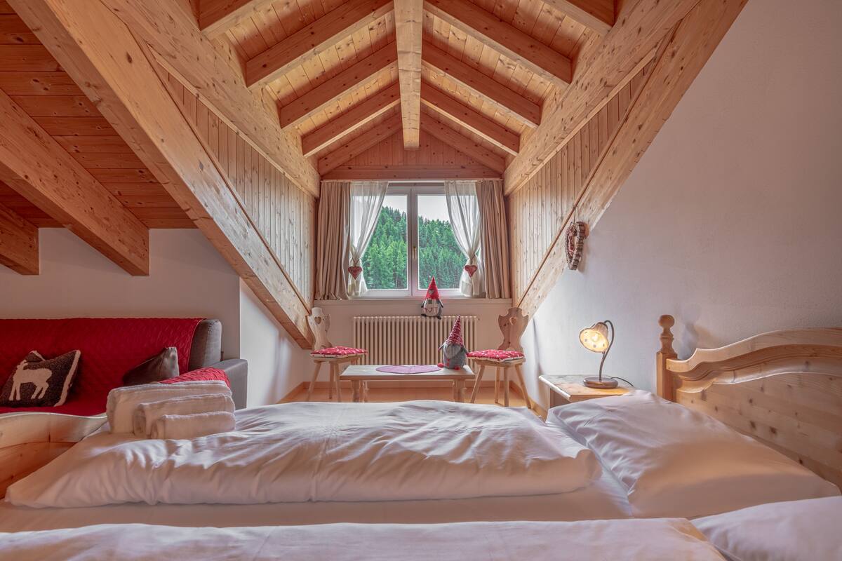 Une chambre confortable avec un plafond en bois, un grand lit, une fenêtre avec des rideaux, une petite table et une table de chevet avec une lampe.