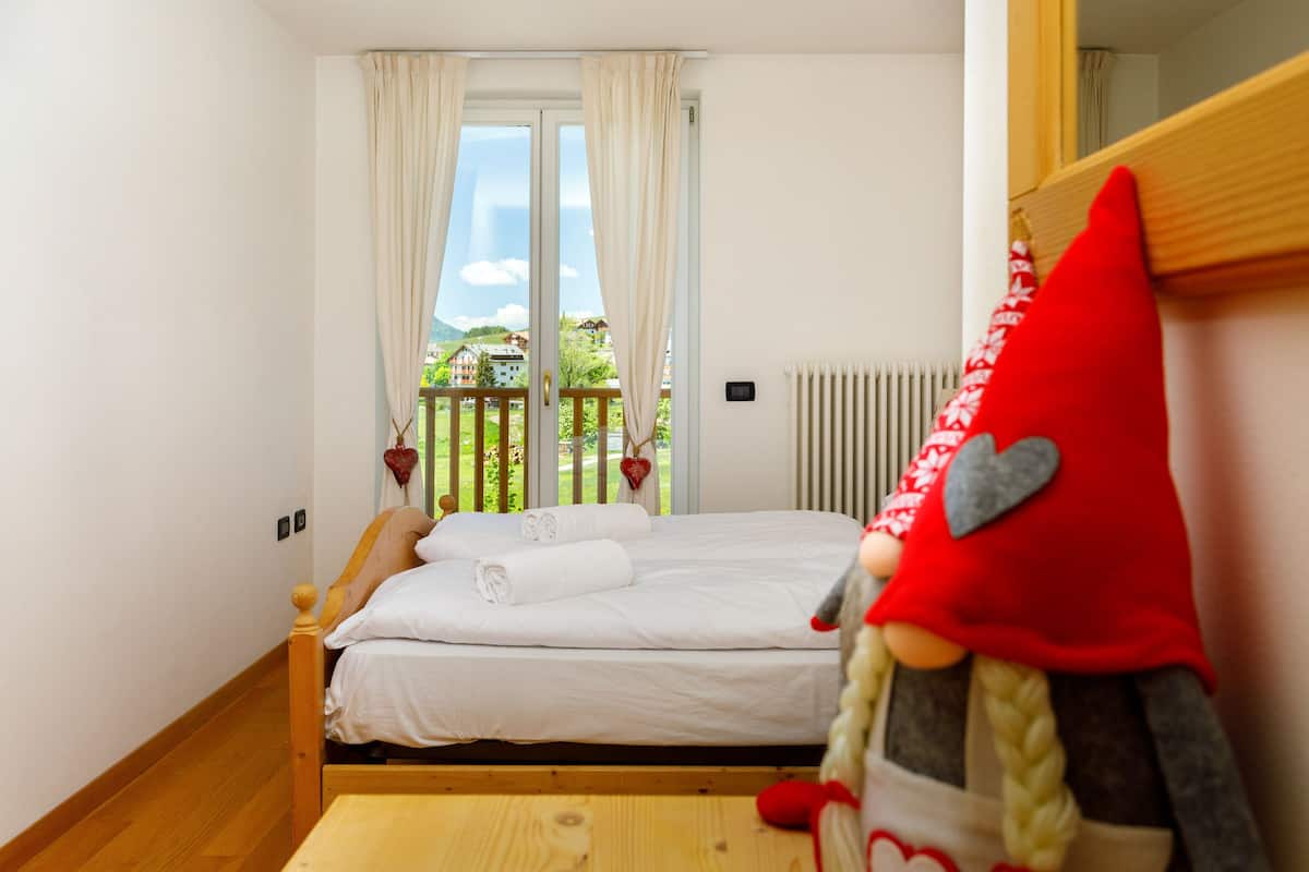 Une chambre avec un lit en bois, du linge de lit blanc, une vue sur un balcon doté d’une balustrade, et un objet décoratif rouge accroché au mur.