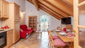 A cozy attic room with wooden beams, a red sofa, a dining table set for two, and a television. - RESIDENCE STELLA (Folgaria)