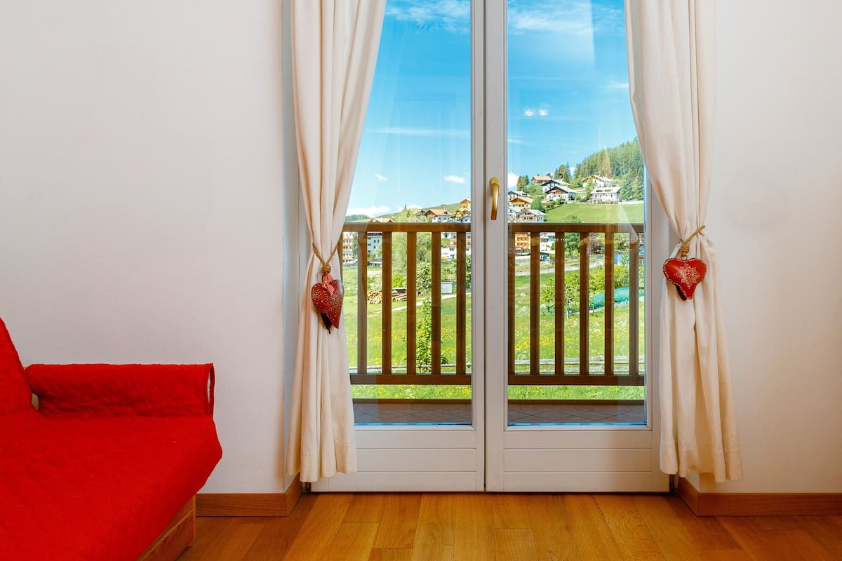Une pièce avec un plancher en bois, un canapé rouge et un balcon donnant sur des maisons et de la verdure.