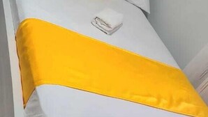 Una cama con ropa de cama blanca y un cojín de acento amarillo, un banco tapizado en gris y un cabecero blanco.