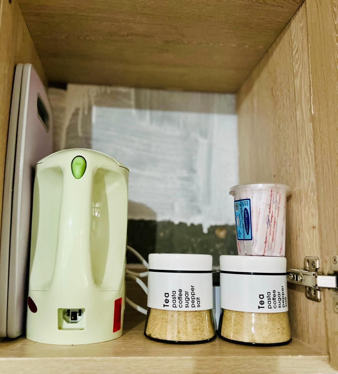 A kitchen with a coffee maker, two spice jars labeled tea, pasta, coffee, sugar, pepper, and salt, and a measuring cup.