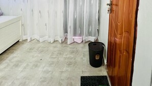 Una habitación con una cama, una puerta de madera, una ventana con cortinas y un felpudo con pantuflas verdes.