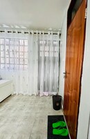 A room with a bed, a wooden door, a window with curtains, and a mat with green slippers.