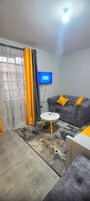 A living room with a grey sofa, yellow cushions, a small white table, and a television on the wall.
