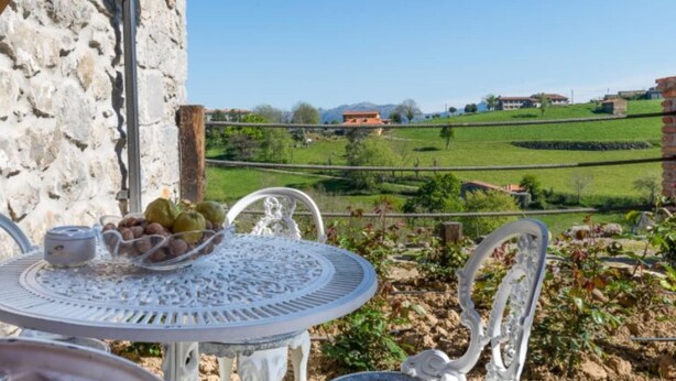 A table with a basket of fruits and a pitcher, set against a stone wall with a view of a green landscape and buildings in the distance. - Apartment - 2 Bedrooms - 8951 (La Aparecida, Cantabria)