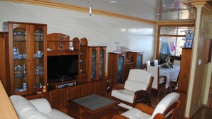 Ein geräumiges Wohnzimmer mit großem Fernseher, Holzschränken, weißen Sofas und einer Essecke mit Tisch und Stühlen.