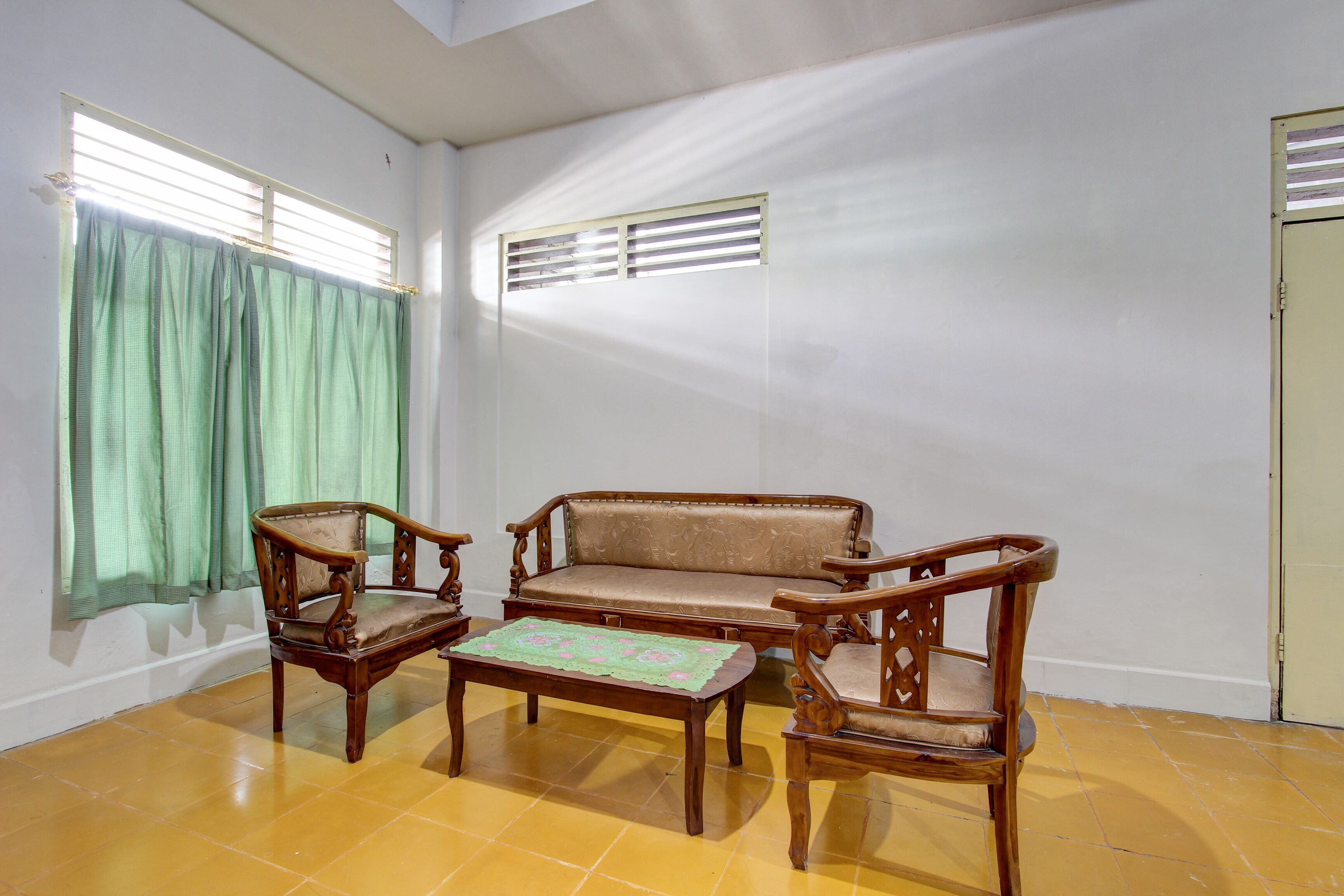 A room with a wooden sofa, two wooden chairs, a small table with a placemat, a window with blinds, and a door.