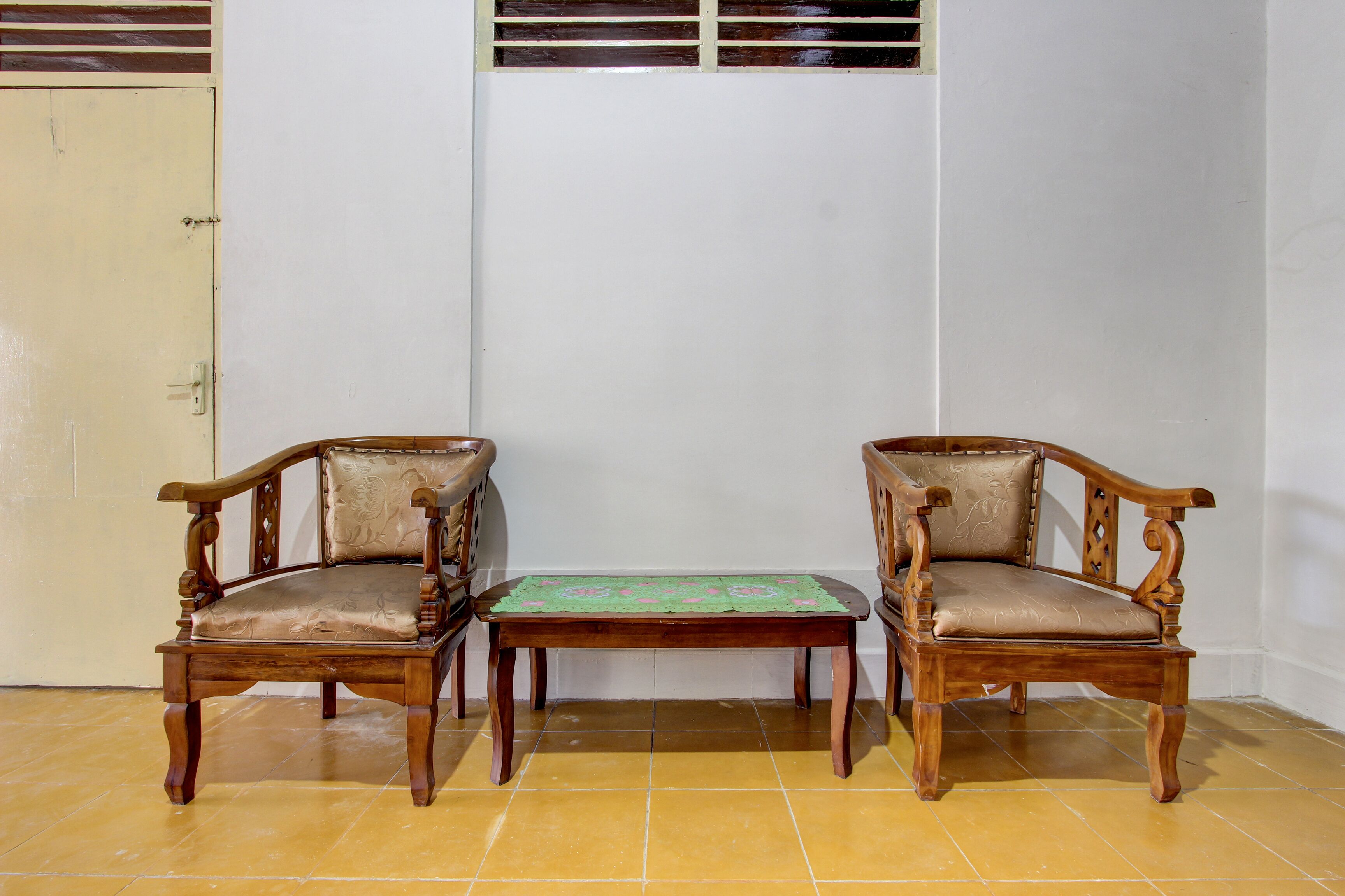 Two wooden chairs with cushioned seats and a small table with a green tablecloth in front of a white wall.