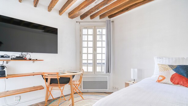 Une chambre à coucher avec un lit, un téléviseur fixé au mur, un bureau avec une chaise et une fenêtre avec des rideaux.
