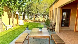 Een terras buiten met een houten tafel en bankjes, omringd door een verzorgd gazon en bomen.