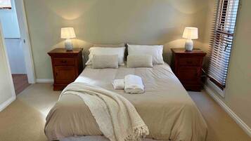 Un lit bien fait, avec une couverture et des oreillers de couleur beige, deux tables de chevet avec des lampes et une fenêtre avec des stores.