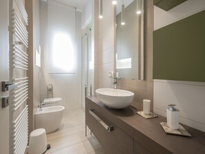 A modern bathroom with a large white sink, a bidet, and a toilet. There is a mirror above the sink and a towel rack to the left.