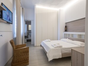 A modern bedroom with a bed, a television, a wicker chair, and a bathroom visible through an open door.