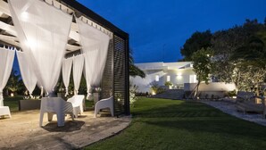 Um pátio moderno ao ar livre, com móveis brancos, uma pérgola com cortinas brancas e um gramado bem cuidado.