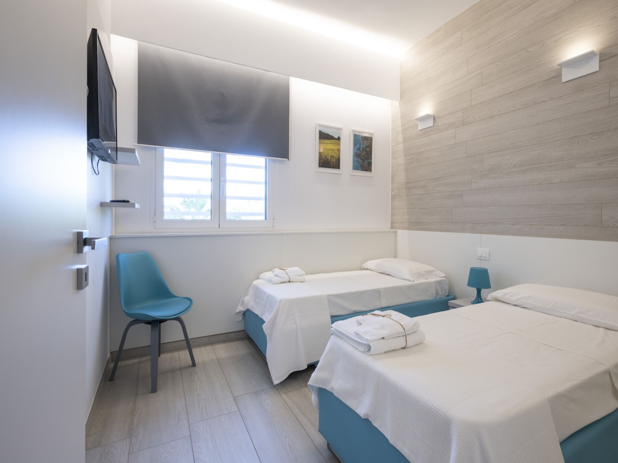 A modern hotel room with two single beds, a blue chair, a flat-screen TV, and a window with blinds.