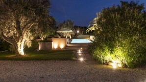 Espaço ao ar livre iluminado à noite, com piscina, caminho e árvores.
