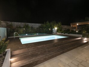 Ein rechteckiger Pool mit einer Holzterrasse und Treppen, umgeben von einer niedrigen Mauer und Grünpflanzen.