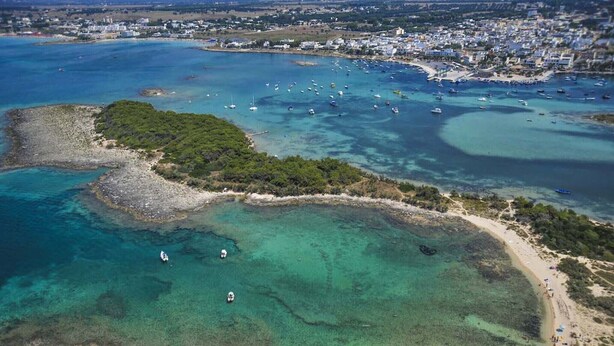 An aerial view of a coastal area with clear turquoise waters, a sandy beach, and a small town in the background.